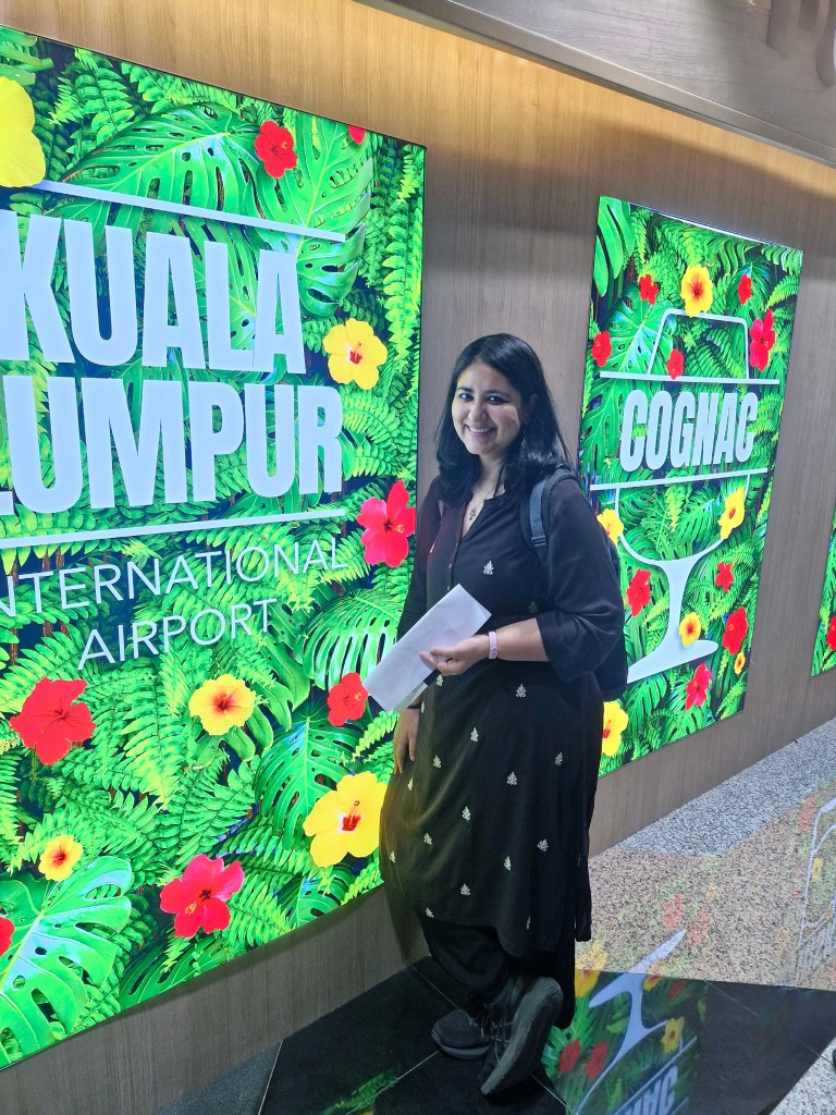 A woman in traditional attire poses in front of vibrant signage featuring tropical plants and flowers that reads 'KUALA LUMPUR INTERNATIONAL AIRPORT' and 'COGNAC'.