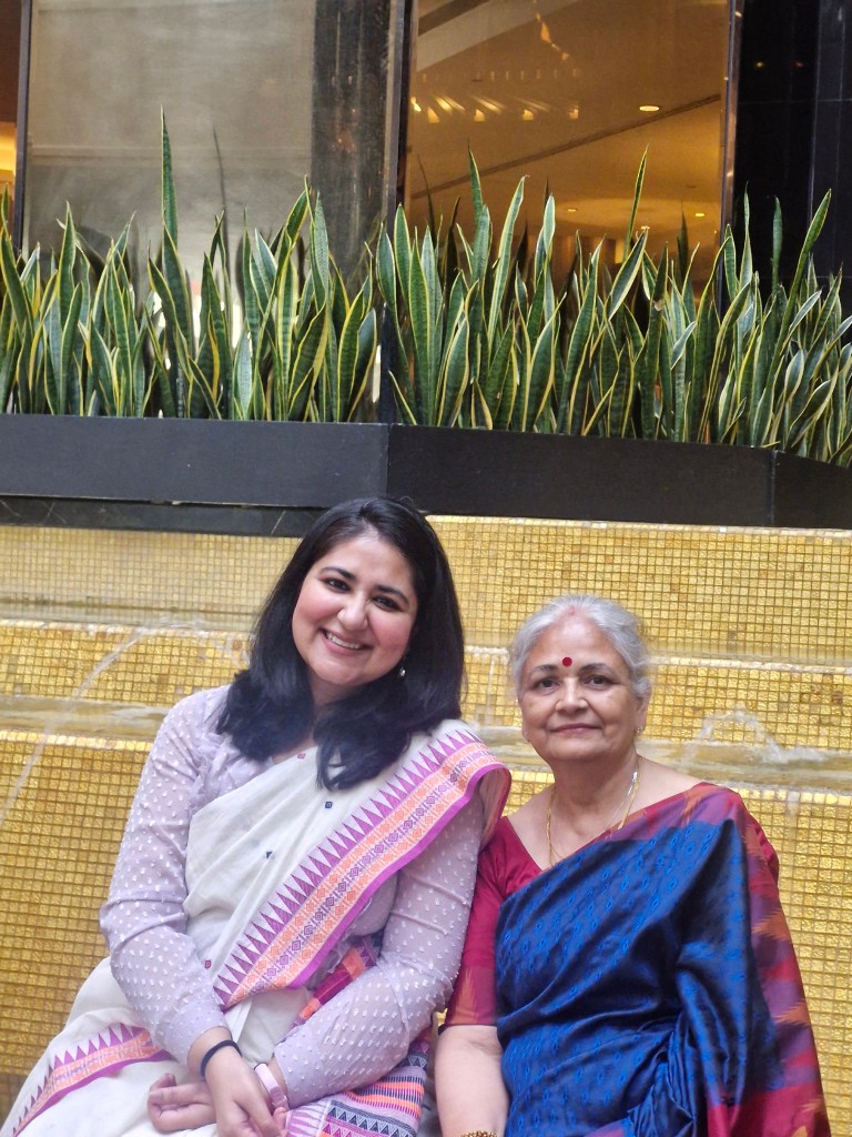 Two women sitting together, smiling. The young woman is wearing a white ethnic outfit with a colourful border, while the older woman is in a blue sari with a red blouse. They are seated against a backdrop of green plants and a golden tiled surface.