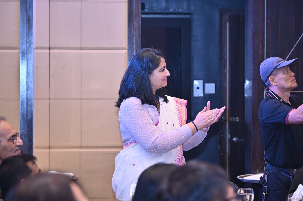 A woman in traditional attire is standing and applauding, while a man in a cap appears to be adjusting equipment in the background.