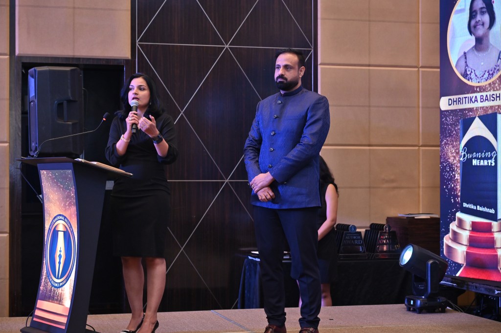 A woman in a black dress speaking at a podium with a microphone, while a man in a blue suit stands beside her. A backdrop features an award and a photo of a girl named Dhritika Baishnab.