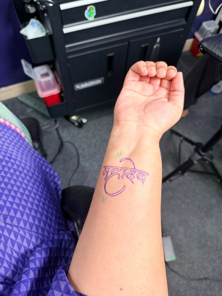 A close-up of a forearm with a newly inked tattoo featuring the word 'महा' in a stylised script, surrounded by small green accents, set against a background of a tattoo studio.