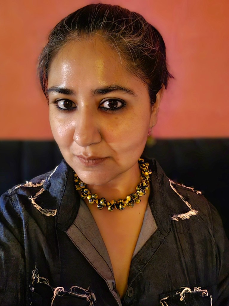 A close-up portrait of a woman with dark hair pulled back, wearing a grey shirt with white patterns and a vibrant yellow and black necklace, against a warm orange background.